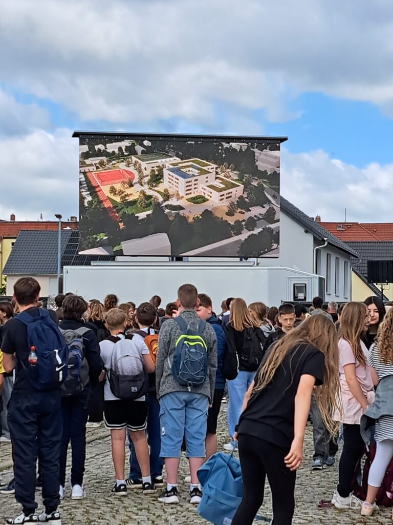 Schülerinnen und Schüler auf einem großen Platz vor einer Leinwand mit Visualisierung des künftigen Bildungscampus Naumburg