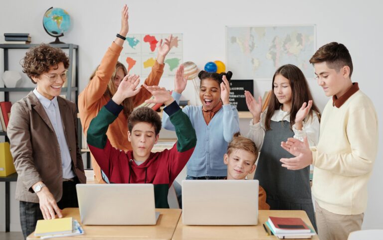 Eine erwachsene Person mit sechs Kindern und Jugendlichen steht neben zwei Tischen. Die Gruppe schaut auf zwei Laptops und klatscht und jubelt.